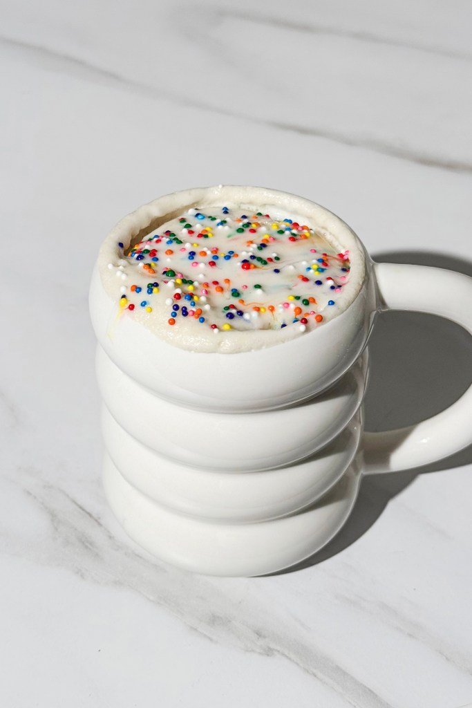 protein dunkaroo mug cake with vanilla icing and rainbow sprinkles in a white mug
